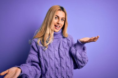 Güzel, sarışın bir kadın mor arka plan üzerine balıkçı yaka kazak giyiyor. Gülümsüyor, kollarını açıp gülümsüyor. Hoş geldin, olumlu ve kendinden emin selamlar.