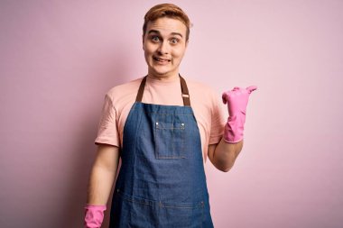 Genç, kızıl saçlı, ev işi yapan, önlük takan, gülümseyen ve baş parmağıyla yüzünü gösteren yakışıklı bir temizlikçi..