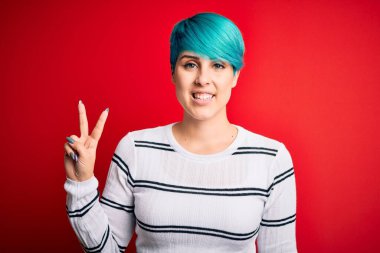 Mavi saçlı, kırmızı arka planda çizgili süveter giyen, kameraya gülümseyen ve zafer işareti yapan güzel bir kadın. İki numara..