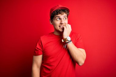 Kıvırcık saçlı genç teslimatçı, izole edilmiş kırmızı arka planda duran şapkalı gergin ve gergin görünüyor ve elleri ağızdan tırnak yiyor. Endişe sorunu.