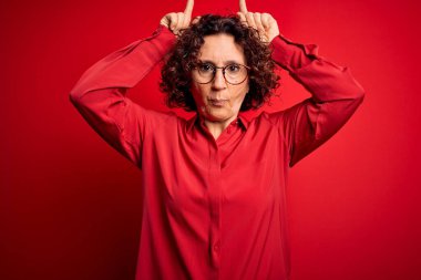 Orta yaşlı güzel kıvırcık saçlı kadın kırmızı arka planda günlük gömlek ve gözlük takıyor ve boynuz gibi parmağıyla komik hareketler yapıyor.