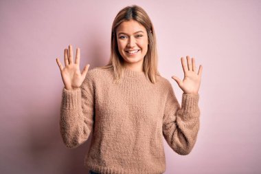 Genç, güzel, sarışın bir kadın pembe yünlü süveter giyiyor. İzole edilmiş arka planda on numaralı parmaklarıyla işaret ediyor. Kendinden emin ve mutlu bir şekilde gülümsüyor..