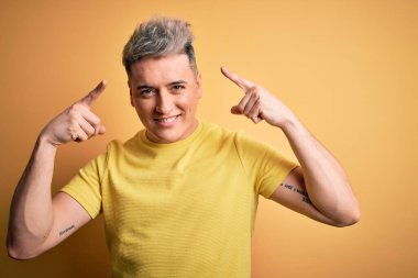 Sarı izole edilmiş arka plan üzerinde sarı tişörtlü, genç ve modern bir adam iki eliyle başını gösteren gülümseme, harika bir fikir ya da düşünce, iyi bir hafıza.