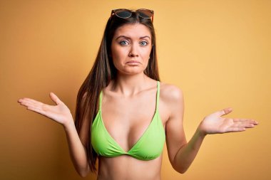 Tatilde mavi gözlü, bikini ve güneş gözlüğü takan, kafası karışmış, elleri ve kolları havada genç ve güzel bir kadın. Şüphe kavramı.