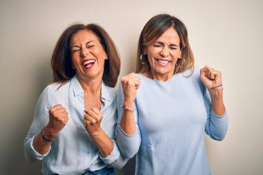 Orta yaşlı, güzel iki kız kardeş izole edilmiş beyaz arka planda duruyorlar. Başarı için heyecanlılar, kolları havada ve gözleri kapalı zaferi kutluyorlar. Kazanan konsept.