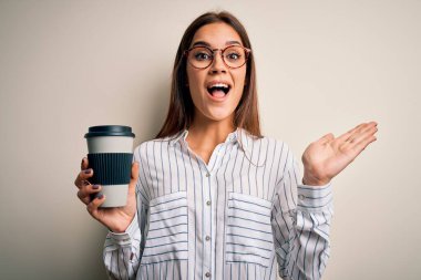 Genç, güzel esmer kadın beyaz arka planda bir fincan kahve içerken mutlu ve heyecanlı, kazanan ifade zaferi büyük bir gülümsemeyle kutluyor ve ellerini kaldırıyor.