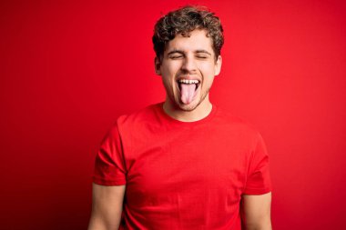 Sarışın, kıvırcık saçlı, kırmızı arka planda sıradan tişörtler giyen, mutlu bir ifadeyle dilini dışarı çıkaran genç, yakışıklı bir adam. Duygu kavramı.