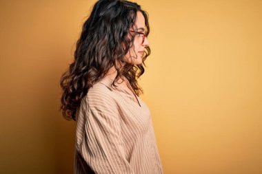 Kıvırcık saçlı, çizgili tişörtlü, sarı arka planda gözlüklü, rahat profil pozu veren, doğal yüzlü ve kendinden emin gülümseyen güzel bir kadın..