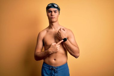 Mayolu ve yüzme şapkalı genç yakışıklı adam izole edilmiş sarı arka planda. Aceleyle saati, sabırsızlığı ve rahat bir ifadeyle kameraya bakıyor.