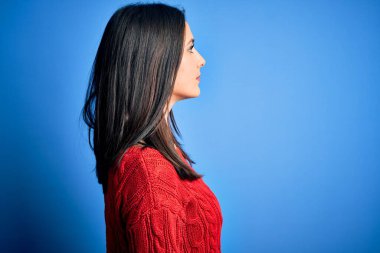 Mavi gözlü genç esmer kadın, izole edilmiş arka planda kırmızı bir süveter giyiyor, yan yana bakıyor, rahat bir görünüm sergiliyor, kendine güvenen bir gülümseme ile doğal bir poz veriyor..