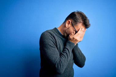 Mavi arka planda duran sıradan süveter ve gözlük takan genç yakışıklı bir adam. Ağlarken yüzünü ellerle kapatan üzgün bir ifade. Depresyon kavramı.