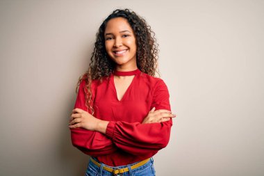 Kıvırcık saçlı, beyaz arka planda duran, kollarını kavuşturup kameraya bakan sıradan tişörtlü güzel bir kadın. Pozitif kişi.