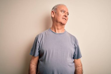 Orta yaşlı, beyaz tişörtlü yakışıklı bir adam izole edilmiş beyaz arka planda duruyor yüzünde gülümsemeyle, doğal bir ifadeyle yan yana bakıyor. Kendinden emin gülen.