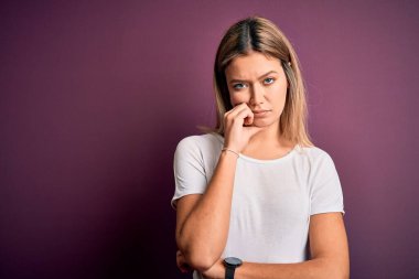 Genç, güzel, sarışın bir kadın mor izole edilmiş arka plan üzerine günlük beyaz tişört giyip kollarını kavuşturarak depresyondan sıkılmış ve yorgun görünüyor..