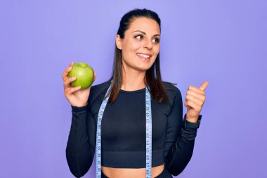 Güzel esmer sporcu kadın mezura kullanarak mor arka planda elma meyvesi yiyor başparmağıyla yan tarafa bakıyor ve ağzı açık gülümsüyor.