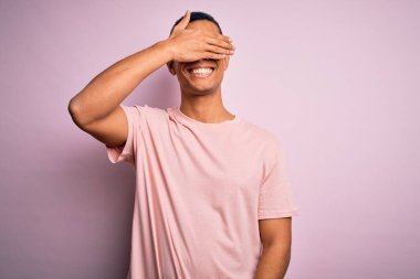 Yakışıklı Afro-Amerikalı adam pembe arka planda günlük tişört ve gözlük takıyor. Gülümsüyor ve yüzünde şaşkınlıktan gözleri kapayarak gülüyor. Kör kavram.