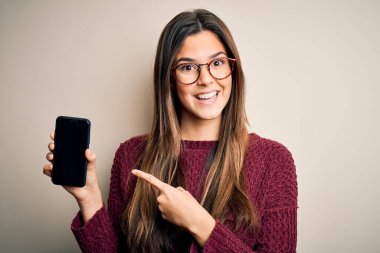 Gözlük takan genç ve güzel bir kız izole edilmiş beyaz arka planda akıllı telefon gösteriyor. Elle ve parmakla işaret ederek çok mutlu.