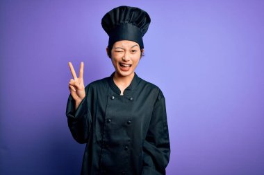 Genç, güzel Çinli aşçı kadın aşçı üniforması ve mor arka planda şapka takıyor. Kameraya gülümsüyor ve parmaklarıyla zafer işareti yapıyor. İki numara..