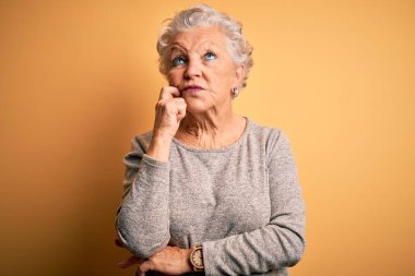 Üst düzey güzel bir kadın, günlük tişört giyer, izole edilmiş sarı arka plan üzerinde durur, elleri çenesinde soru sorarken, düşünceli bir ifade takınır. Düşünceli bir yüzle gülümsüyor. Şüphe kavramı.