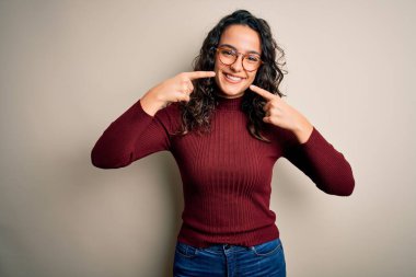 Kıvırcık saçlı, günlük süveter giyen ve beyaz arka planda gözlüklü neşeli gülümseyen ve parmaklarıyla ve ağzıyla işaret eden güzel bir kadın. Diş sağlığı konsepti.