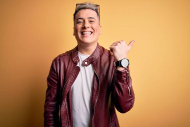 Genç, yakışıklı, deri ceket giyen, sarı arka planda güneş gözlüğü takan mutlu yüzüyle gülümseyen ve baş parmağıyla yüzünü gösteren modern bir adam..