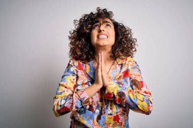 Genç, kıvırcık, güzel, kıvırcık Arap kadın, çiçek rengi gömlek giyiyor, beyaz arka planda duruyor, yalvarıyor ve dua ediyor. Yüzünde umut ifadesi var, çok duygusal ve endişeli. Yalvarıyorum..