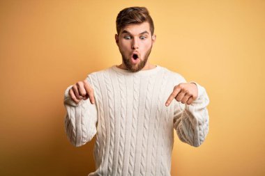 Sarışın, sakallı, mavi gözlü, sarı arka planda beyaz süveter giyen, reklamını yapan, şaşırmış yüzünü ve ağzını açan genç bir adam.