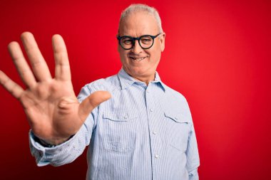 Orta yaşlı, yakışıklı, beyaz gömlekli, kırmızı arka planda gözlüklü, kendinden emin ve mutlu bir şekilde gülümseyen, beş numarayı işaret eden bir adam..