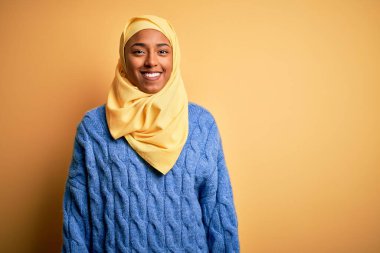 Genç Afrikalı Afro kadın, izole edilmiş sarı arka plan üzerine Müslüman başörtüsü takıyor. Yüzünde mutlu ve havalı bir gülümseme var. Şanslı kişi..