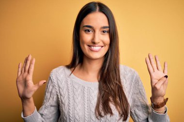 Sarı arka planda beyaz süveter giyen genç, güzel esmer kadın dokuz numarayı gösteriyor ve gülümsüyor, kendinden emin ve mutlu..