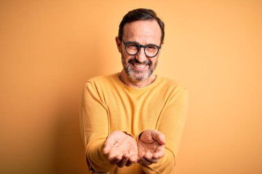Orta yaşlı, ağarmış, günlük süveter ve gözlük takan, izole edilmiş sarı arka planda gülümseyen, avuç içleriyle gülümseyen, el hareketi yapan bir adam. Tutun ve koruyun