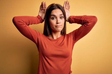 Sarı arka plan üzerinde sıradan tişörtler giyen genç, güzel esmer kadın avuçlarında alaycı ve şüpheci bakışlarla tavşan kulağı hareketi yapıyor. Paskalya tavşanı kavramı.