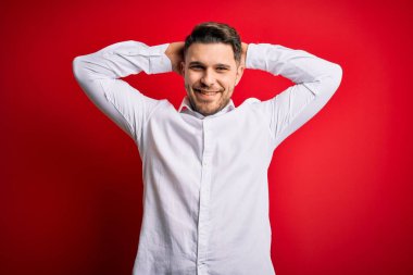 Mavi gözlü, zarif gömlekli genç bir iş adamı kırmızı izole edilmiş arka planda duruyor rahatlıyor ve esniyor, kolları ve elleri başının ve boynunun arkasında mutlu bir şekilde gülümsüyor.