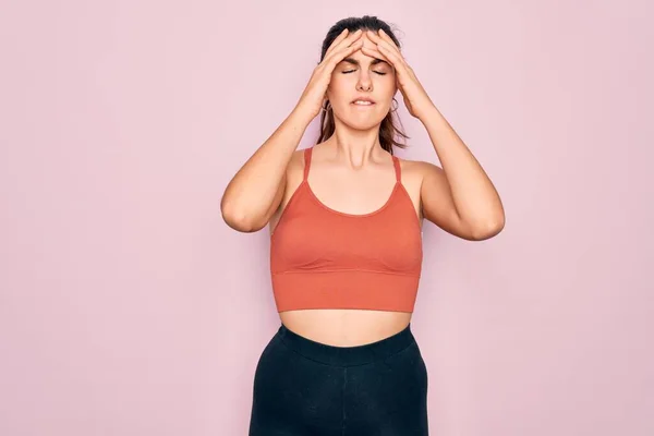 Young beautiful fitness woman wearing sport excersie clothes over pink background suffering from headache desperate and stressed because pain and migraine. Hands on head.