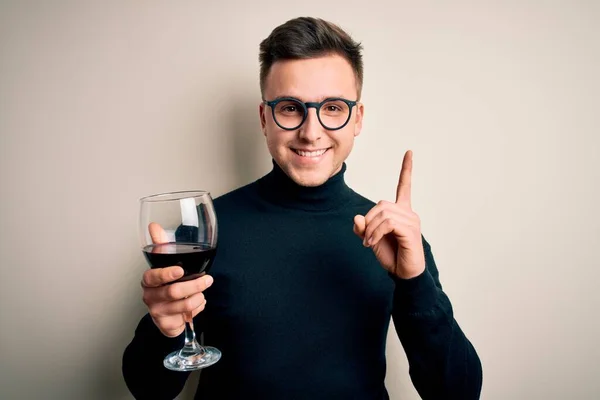 若いですハンサムなCaucasian男飲みますAアルコールガラスの赤ワイン上の隔離された背景驚きでアイデアや質問を指差す指で幸せな顔 ナンバーワン — ストック写真
