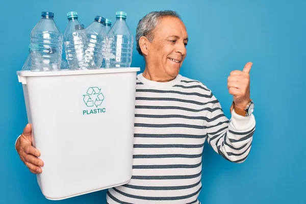 ゴミを持っている高齢者は 青い背景の上にリサイクルし 幸せな顔を笑顔で親指で横に示すためにペットボトルで缶をリサイクル — ストック写真