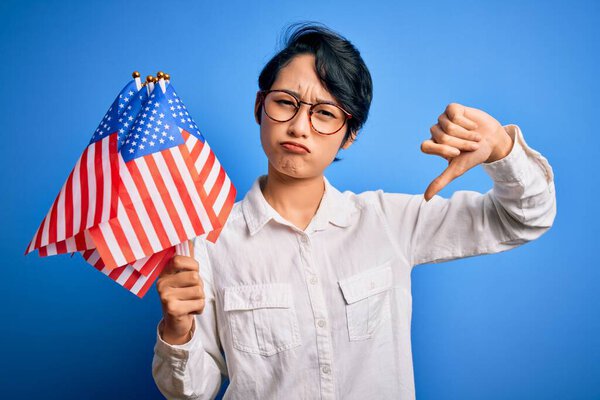 Young beautiful asian patriotic girl holding united states flags celebrating independence day with angry face, negative sign showing dislike with thumbs down, rejection concept