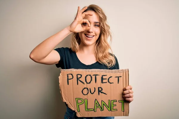 Young activist woman asking for environment holding banner with protect planet message with happy face smiling doing ok sign with hand on eye looking through fingers