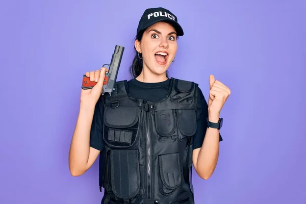 Young police woman wearing security bulletproof vest uniform and holding gun screaming proud and celebrating victory and success very excited, cheering emotion