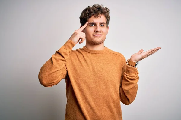 Young blond handsome man with curly hair wearing casual sweater over white background confused and annoyed with open palm showing copy space and pointing finger to forehead. Think about it.