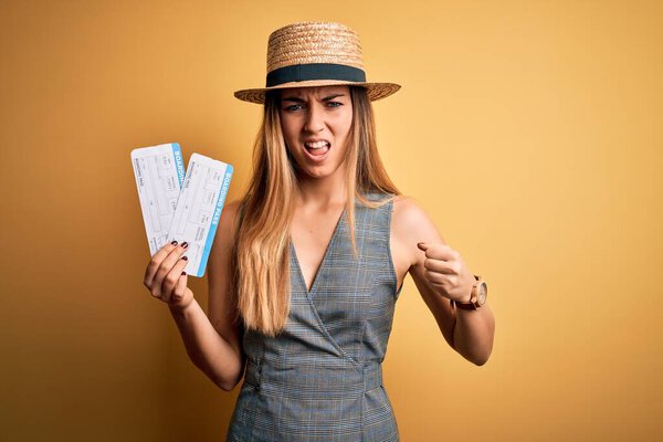 Young blonde tourist woman with blue eyes on vacation wearing hat holding boarding pass annoyed and frustrated shouting with anger, crazy and yelling with raised hand, anger concept