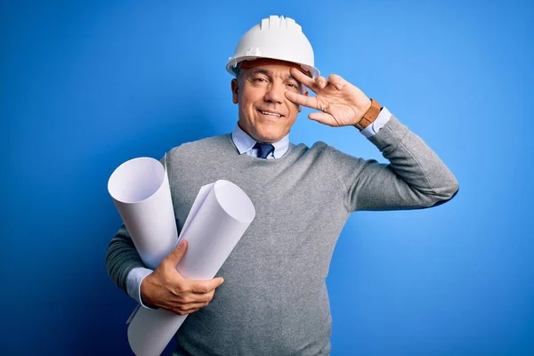 Middle age handsome grey-haired architect man wearing safety helmet holding blueprints Doing peace symbol with fingers over face, smiling cheerful showing victory