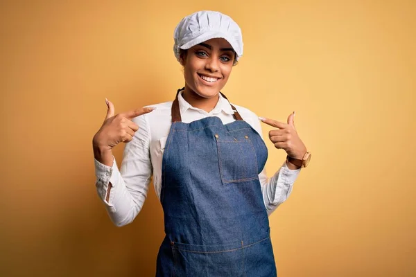 年轻的非洲裔美国面包师女人 头戴围裙 头戴黄帽 面带微笑 带着自信 用手指指着自己 自豪而快乐 — 图库照片