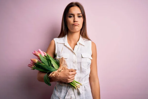 Young beautiful brunette woman holding bouquet of tulips flowers over pink background depressed and worry for distress, crying angry and afraid. Sad expression.