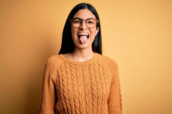 黄色の隔離された背景にメガネをかけている若い美しいヒスパニック系の女性が面白い表情で幸せ舌を突き出しています 感情概念 — ストック写真
