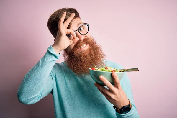 Redhead Irish healthy man with beard eating vegetarian green salad over pink background with happy face smiling doing ok sign with hand on eye looking through fingers