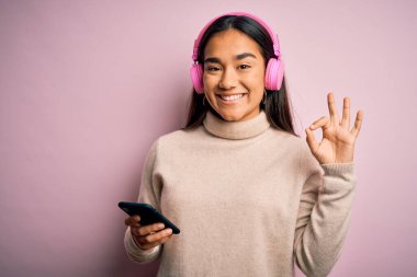Genç, güzel Asyalı bir kadın akıllı telefon ve kulaklık kullanarak müzik dinliyor. Parmaklarıyla imza atıyor. Mükemmel bir sembol.