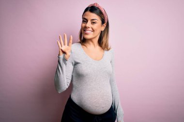 Genç, güzel, esmer bir kadın hamile, pembe arka planda bebek bekliyor. Dört numaralı parmaklarıyla işaret ediyor. Kendinden emin ve mutlu bir şekilde gülümsüyor..