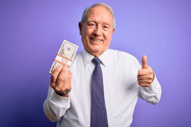 Mor arka planda 50 dolarlık banknotlar tutan yaşlı gri saçlı adam gülümsüyor. Tamam işareti yap, parmaklarınla başparmağını kaldır, mükemmel bir işaret.