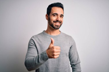Sakallı, sıradan bir süveter giyen, beyaz arka planda duran genç ve yakışıklı bir adam mutlu bir el hareketi yapıyor. Kameraya bakıp başarıyı gösteren ifade onaylanıyor.